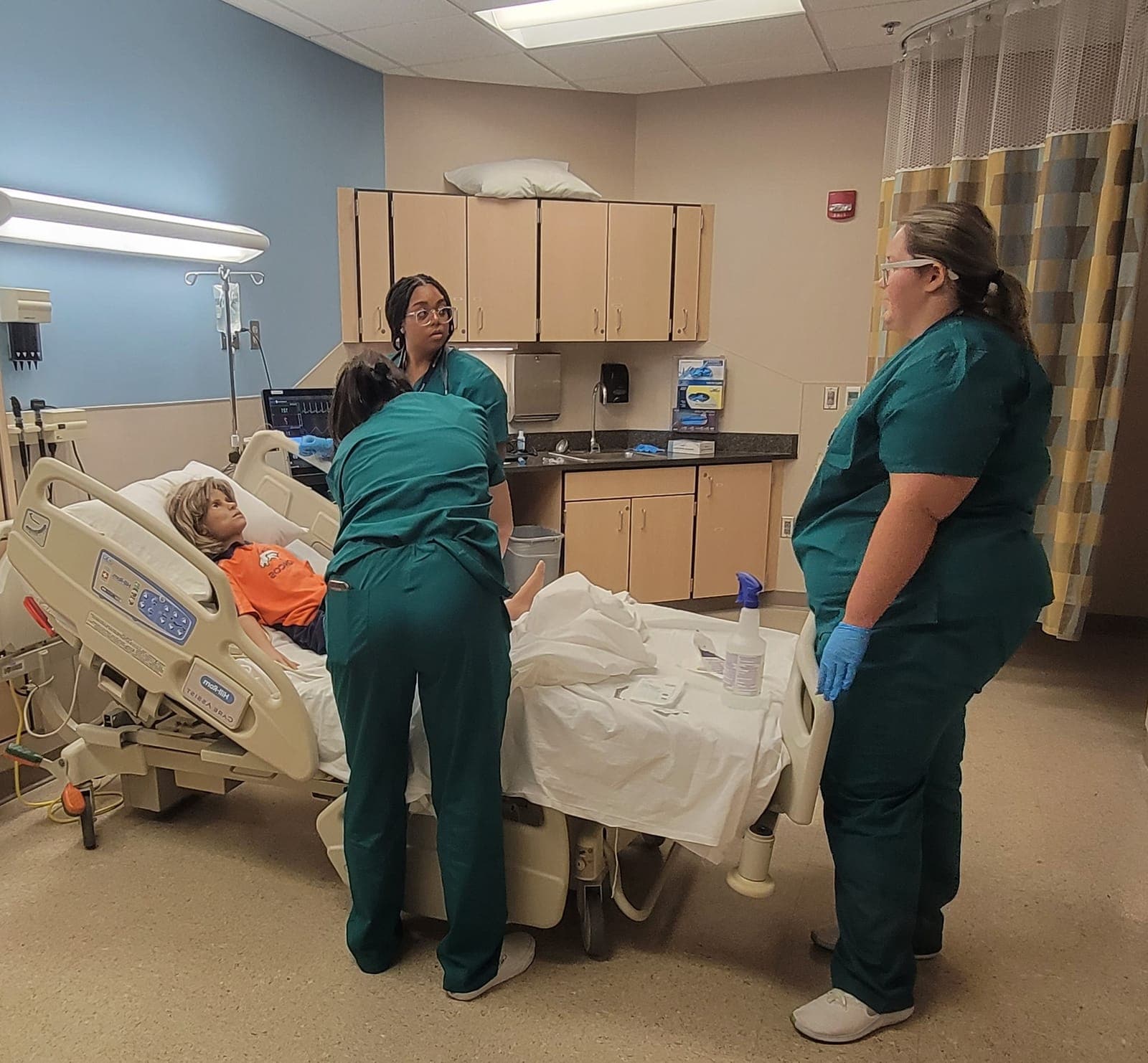 Three students in teal scrubs care for a child patient simulator in a hospital simulation room at HOPE Camp